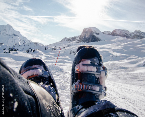 Nahaufnahme von Füßen mit alpinen Skistiefeln und Skipiste und schneebedeckte Berggipfel im Hintergrund photo