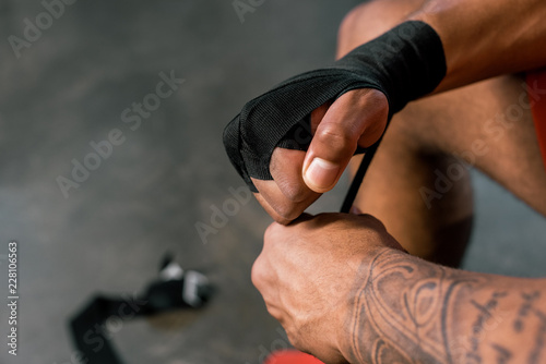 cropped image of tattooed sportsman wrapping hand in boxing bandage at gym