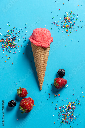 Top view of a pink icecream scoop in a waffle cone with strawberries, black berries, and colorful, rainbow sprinkles on a blue background