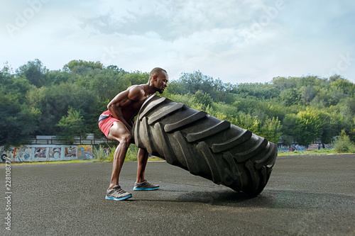 Fotografi Handsome african american muscular man flipping big tire outdoor.