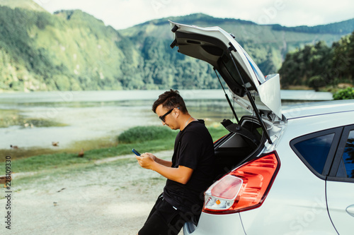 Young travel man in sunglasses sitting in car trunk typing on phone with beauty nature landscape on background. Summer travel.