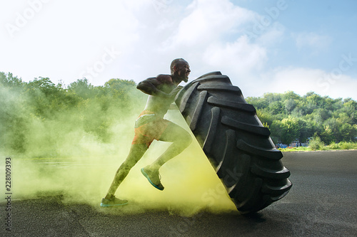 Handsome african american muscular man flipping burning big tire outdoor with smoke