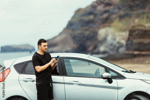Young man use phone for network surfing or check mail white standing in front of the car with ocean rock background. Summer vocation.
