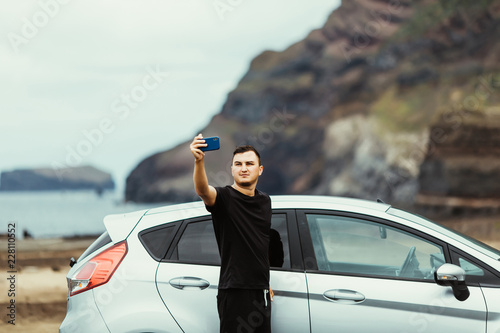 Young man take selfie on the phone near car with ocean view. Car trip. Summer vocation.
