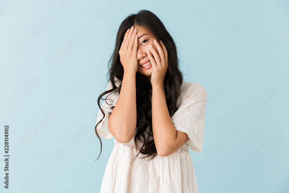 Cute young asian beautiful woman posing isolated over blue background covering face.