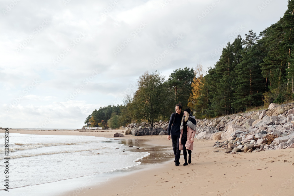 Young couple in love walking on the beach coast. Cold autumn weather, trees in the background. Young man wearing a wool sweater, girl in a pink coat