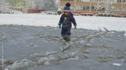 Cute child breaking ice layer of big frozen puddle with boots photo