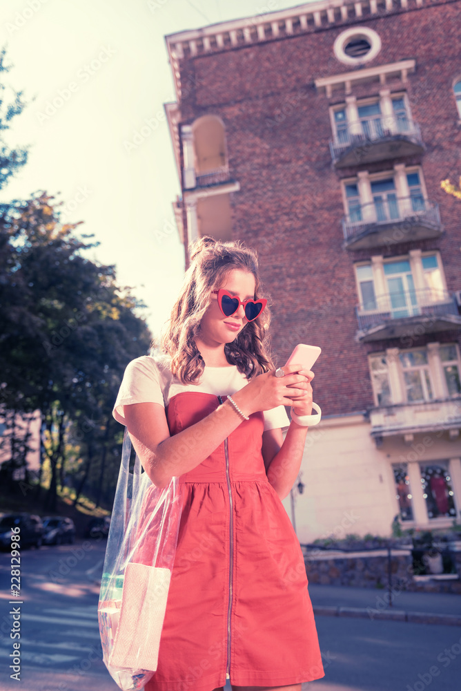 Reading message. Good-looking curly woman wearing red dress and bright sunglasses feeling pleased reading positive message on phone