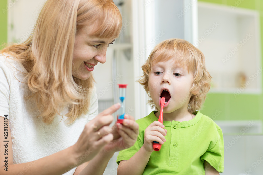 Kid brushing teeth near mirror in bathroom. His mother monitoring accuracy and time of cleaning action with hourglass. Kid brushing teeth near mirror in bathroom. His mother monitoring accuracy and time of cleaning action with hourglass.