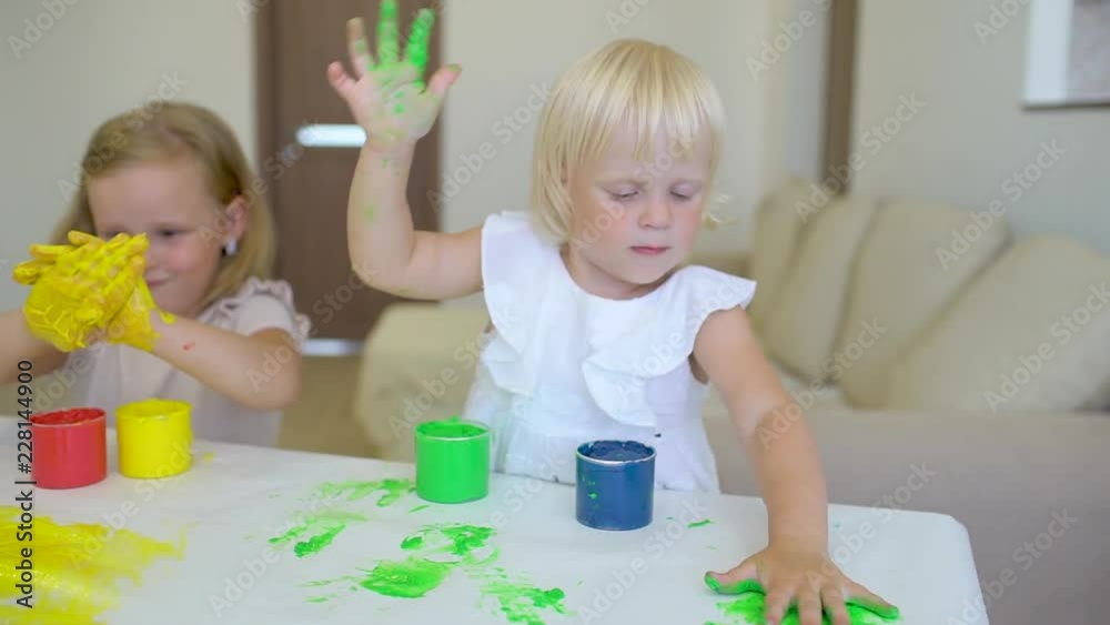 Happy family paints colors. Happy little childs making colorful color handsprint on the white paper. A little cute happy funny girls painting color with smiling mother together. Slow motion Happy family paints colors. Happy little childs making colorful color handsprint on the white paper. A little cute happy funny girls painting color with smiling mother together. Slow motion
