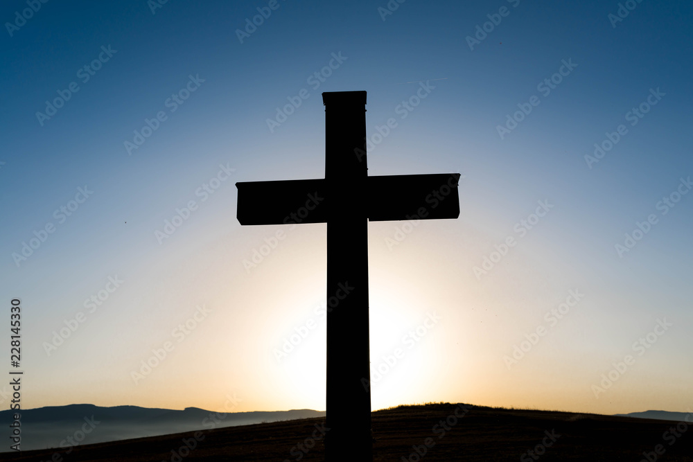 Simple oak wood catholic cross silhouette at sunset, dramatic dark blue sky with the glowing sun.