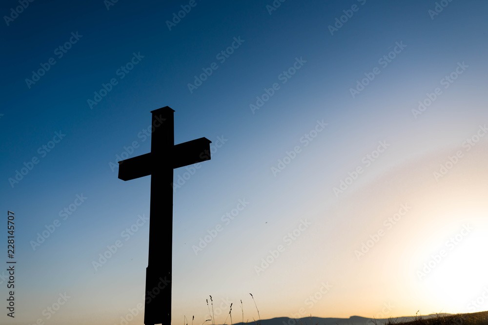 Simple oak wood catholic cross silhouette at sunset, dramatic dark blue sky with the glowing sun. Simple oak wood catholic cross silhouette at sunset, dramatic dark blue sky with the glowing sun.