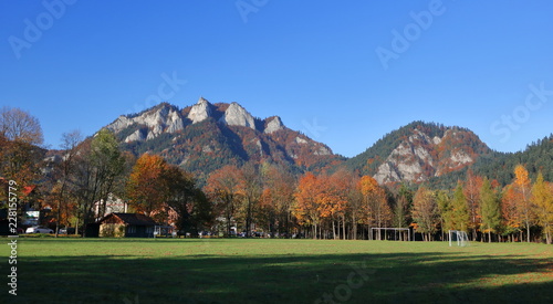 panoramic view at Three Crowns massif in Pieniny Mountains, Poland, from valley in Srmowce Nizne village, field, trees with colorful autumnal foliage, sharp peaks of limestone rocks, clear blue sky.