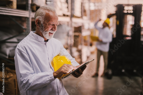 Picture of older male warehouse employee standing in storage and looking at his tablet. Dressed in white coat and yellow helmet. photo