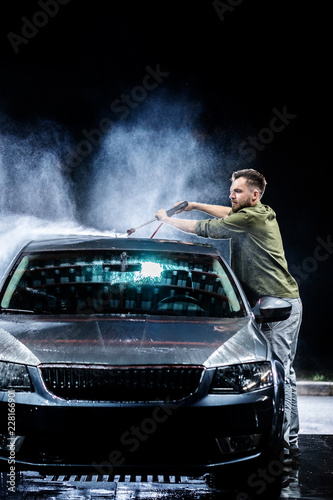 A man with a beard or car washer washes a gray car with a high-pressure washer at night in on the street © vasilkamalov
