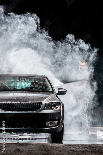 A man with a beard or car washer washes a gray car with a high-pressure washer at night in a shop wash