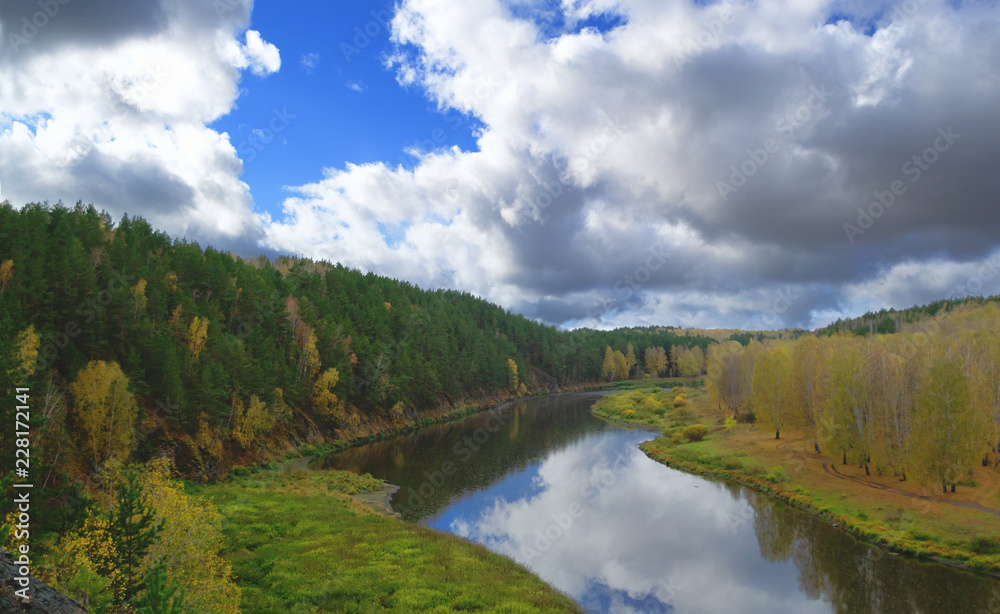 river against autumn forest with yellow and green trees and bright colorful blue sky with clouds