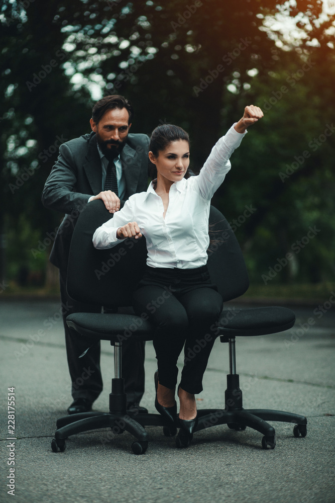 Young woman sitting on two office chairs outside. Man pushing the behind. Making fun after work. Young woman sitting on two office chairs outside. Man pushing the behind. Making fun after work.