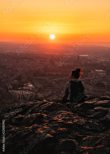 The girl from the back is sitting on a rock of a high mountain watching the sunset over the cit