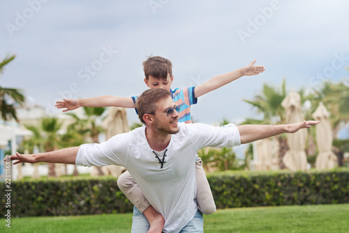 Boy is sitting on his fathers back. They are having fun. Their hands are open wide and they are imitating airplanes.