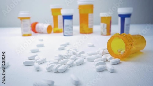Bottle of painkillers falls on a pile of white narcotic pills in slow motion.  Prescription bottles clutter the background. The capsules represent the opioid crisis in America and narcotic addiction.