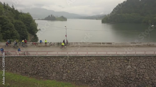 Aerial view, orbit style, of a group of cyclists standing over the dam at Colibita Lake while one cyclist peddles along, during Tura Cu Copaci race. photo