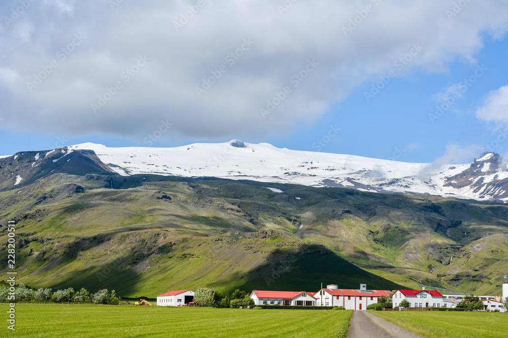 Eyjafjallajokull volcano in Iceland against blue summer sky with clouds. Farm Thorvaldseyri and path visible.