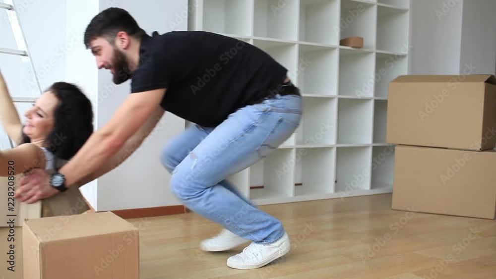 Real Eatate. Happy Young Couple Moving House Together With The Pretty Young Woman Sitting Inside a Brown Cardboard Box With Her Husband Crouching Alongside Smiling Real Eatate. Happy Young Couple Moving House Together With The Pretty Young Woman Sitting Inside a Brown Cardboard Box With Her Husband Crouching Alongside Smiling