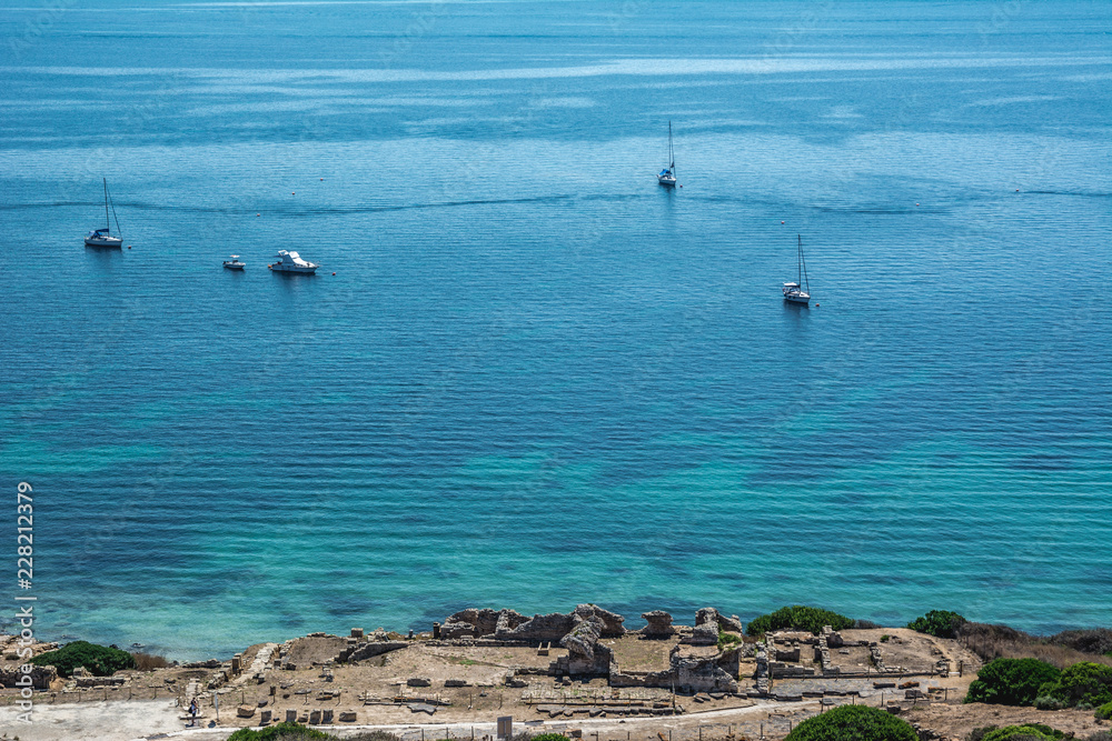 Beautiful turquoise sea with yachts and small part of Corinthian ruins of Tharros village in Sinis peninsula, Cabras, Oristano province, Sardinia, Italy. Beautiful turquoise sea with yachts and small part of Corinthian ruins of Tharros village in Sinis peninsula, Cabras, Oristano province, Sardinia, Italy.