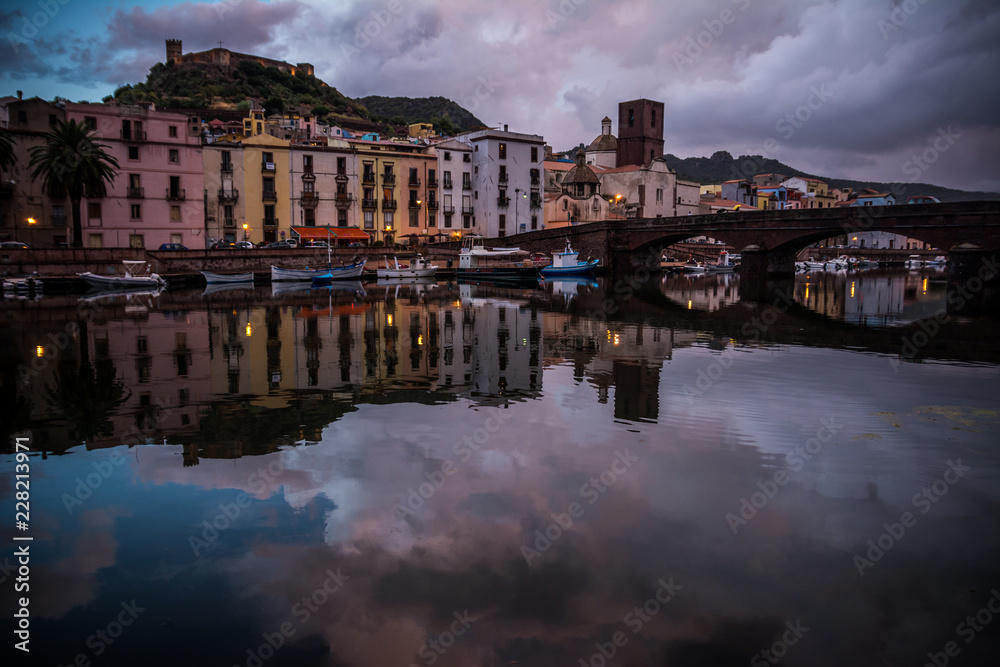Obraz premium Night city view of Bosa, Sardinia, Italy. Colorful houses and beautiful clouds reflecting in the water after rain at dusk.