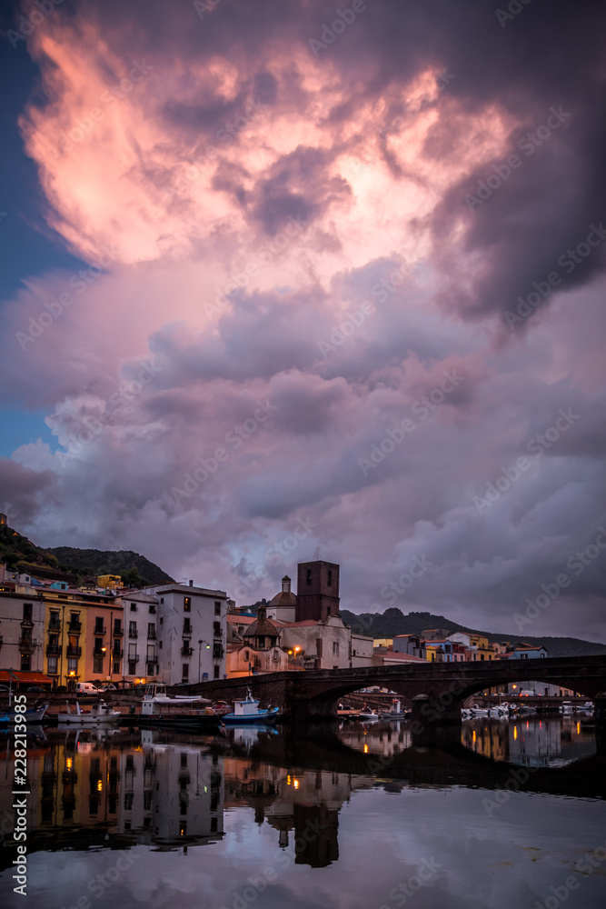 Night city view of Bosa, Sardinia, Italy. Colorful houses and beautiful clouds reflecting in the water after rain at dusk.