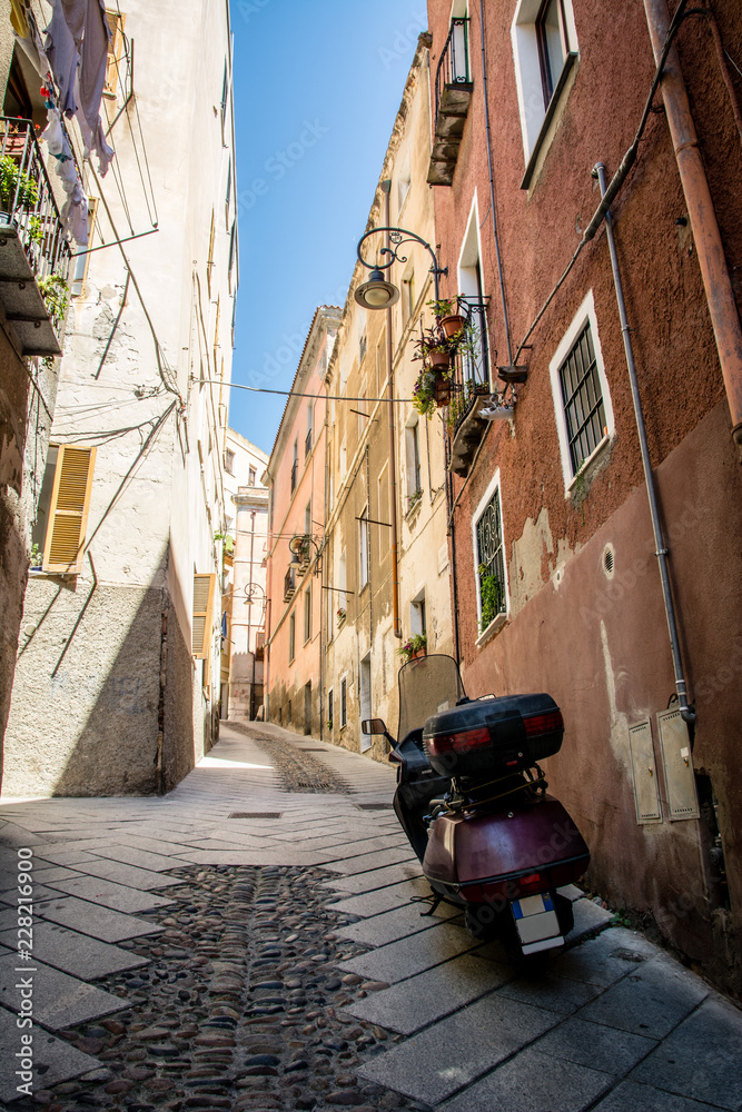 Narrow steep streets of Cagliari in Sardinia, Italy with view up. Beautiful blue sky on a sunny day in the background. Motorcycle parked on sideways. Nobody in the scene.