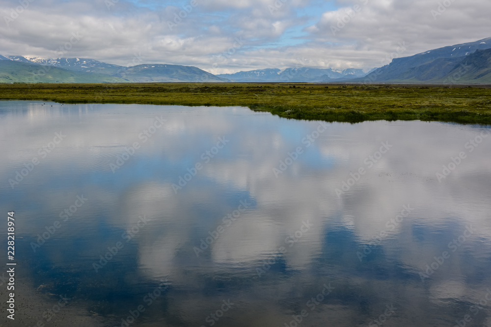 Beautiful cloudy sky reflecting in calm water with fresh green field and mountains in Iceland