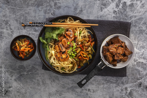 Chinese noodles with mushrooms, fried meat in a pan, vegetables and herbs are on the gray table. Top view