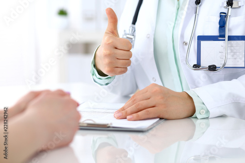 Doctor showing Ok sign with thumb up to patient while sitting at the desk in hospital office, close-up of human hands. Medicine and health care concept