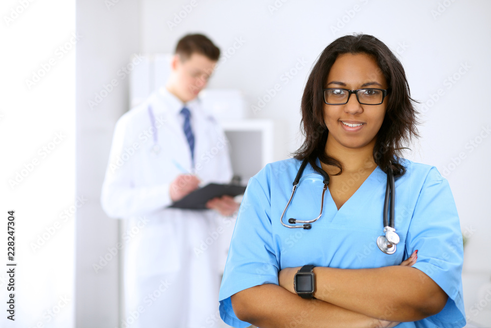 Female african american medical doctor with colleagues in background at hospital. Medicine and health care concept Female african american medical doctor with colleagues in background at hospital. Medicine and health care concept
