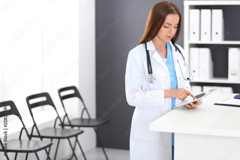Doctor woman at work. Portrait of female physician using digital tablet while standing near reception desk at clinic or emergency hospital. Medicine and healthcare concept Doctor woman at work. Portrait of female physician using digital tablet while standing near reception desk at clinic or emergency hospital. Medicine and healthcare concept