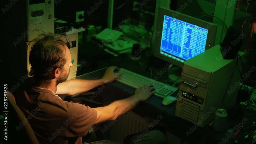 Adult hacker works with old software on retro computers with blue screen and dark green lighting Adult hacker works with old software on retro computers with blue screen and dark green lighting