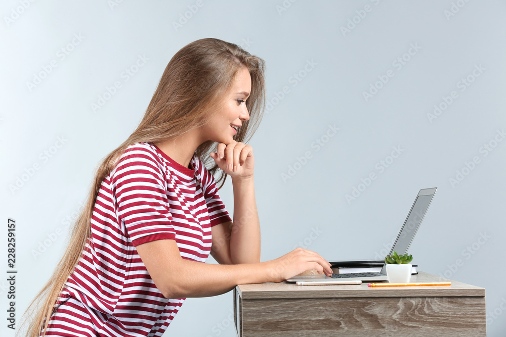 Young woman working with laptop while sitting at table against grey background Young woman working with laptop while sitting at table against grey background