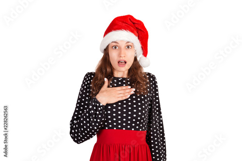 surprised woman in dress holding hands on breast smiling being excited and shocked. emotional girl in santa claus christmas hat isolated on white background. holiday concept