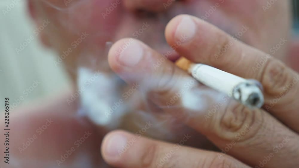 Macro shot of a lit cigarette with grey ash in the mouth of a tranquil man who breathes out a stream of thick smoke in summer in slow motion Macro shot of a lit cigarette with grey ash in the mouth of a tranquil man who breathes out a stream of thick smoke in summer in slow motion