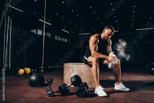 handsome athletic man sitting on cube with gym equipment around and clapping hands with talc before workout