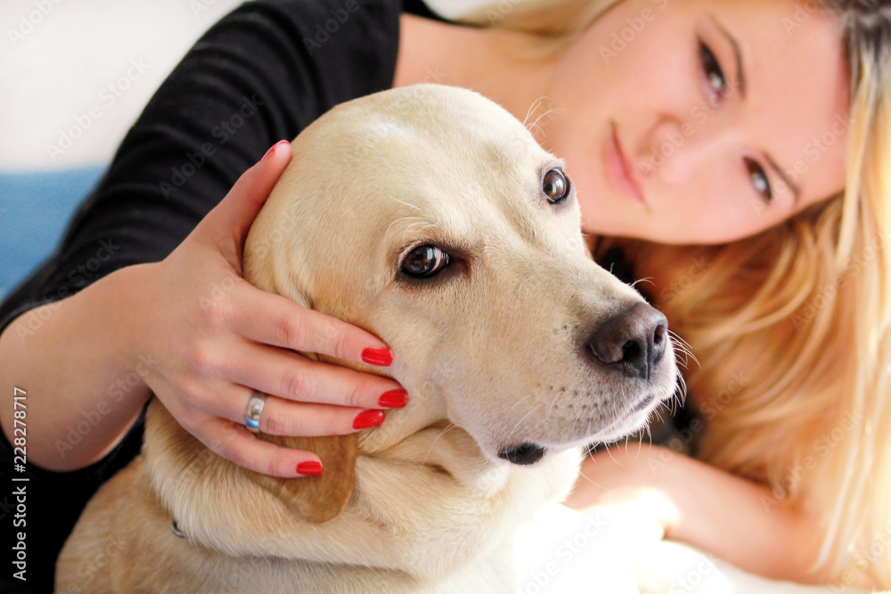 Woman with dog is resting in bed at home, relaxing in bedroom. Girl is petting with her dog. Portrait of cute yellow labrador retriever and her owner, enjoying on blue bed, posing in front of camera.