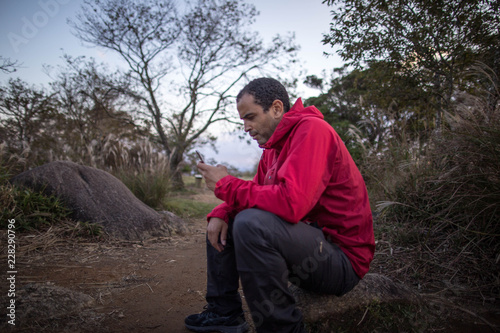 Man in red jacket using phone in left hand beside dirt trail in nature © Osaze