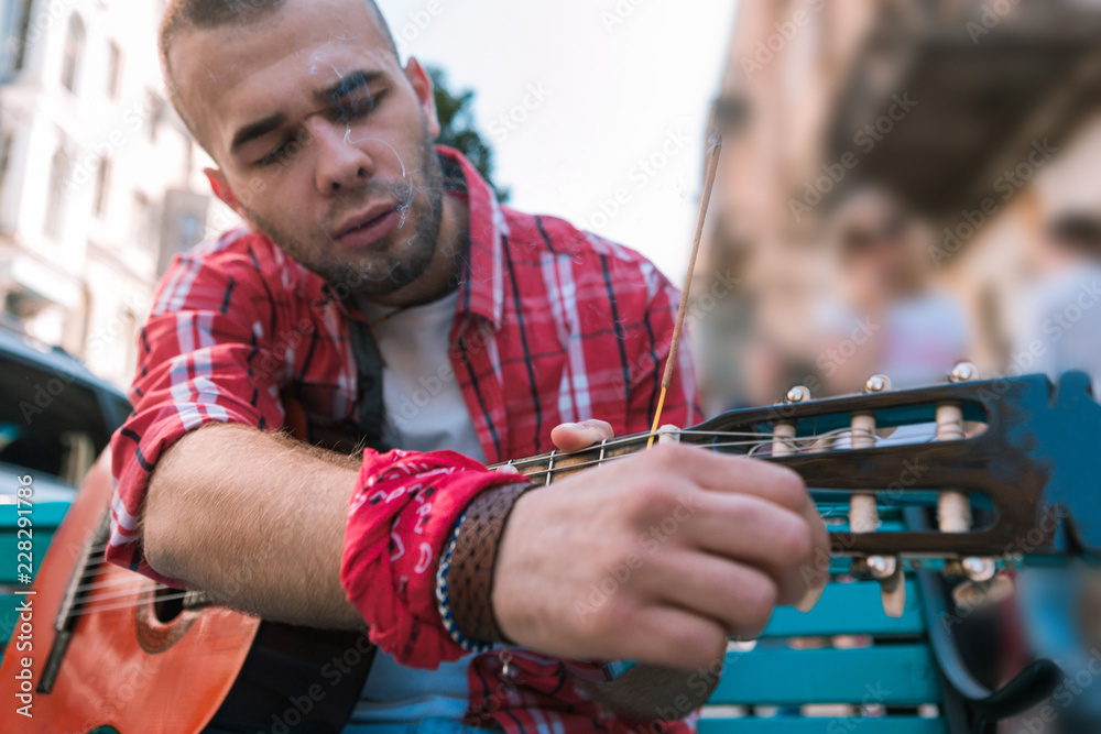 Fine tune. Low angle of serious street musician adjusting guitar and posing on blurred background