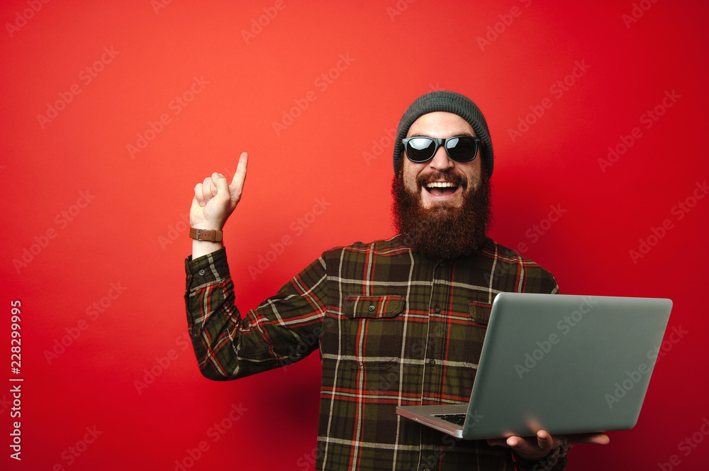 Happy man with beard holding laptop and pointing up over red background, copyspace Happy man with beard holding laptop and pointing up over red background, copyspace