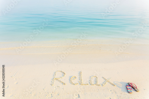Fototapeta Naklejka Na Ścianę i Meble -  The Word Relax Written in the Sand on a Beach with flup flops at morning sea background