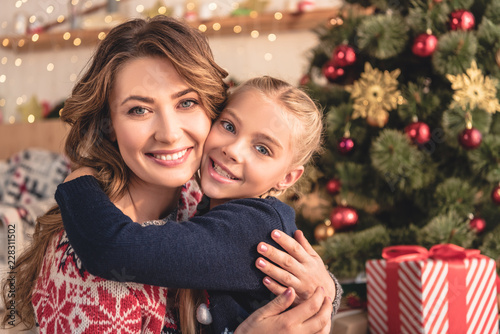 smiling mother and daughter hugging and looking at camera near christmas tree at home
