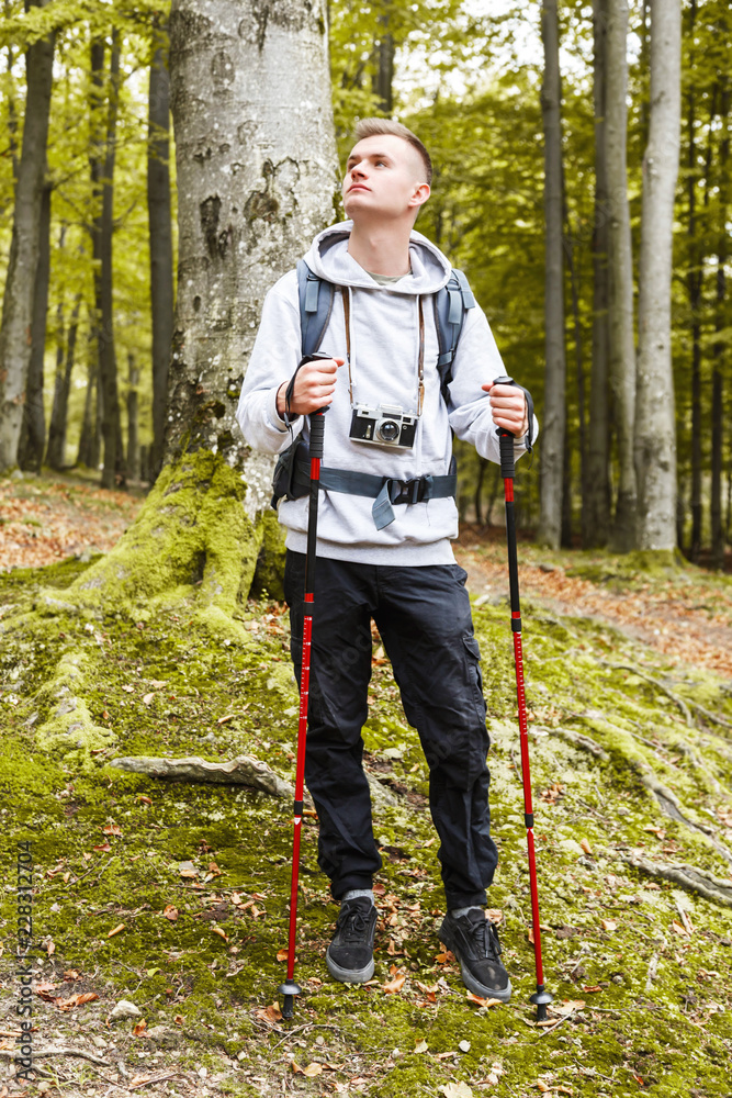 Fototapeta premium Young blonde attractive man with trekking poles standing in the forest, he searching some places and enjoying nature, trekking, recreation and healthy lifestyle concept