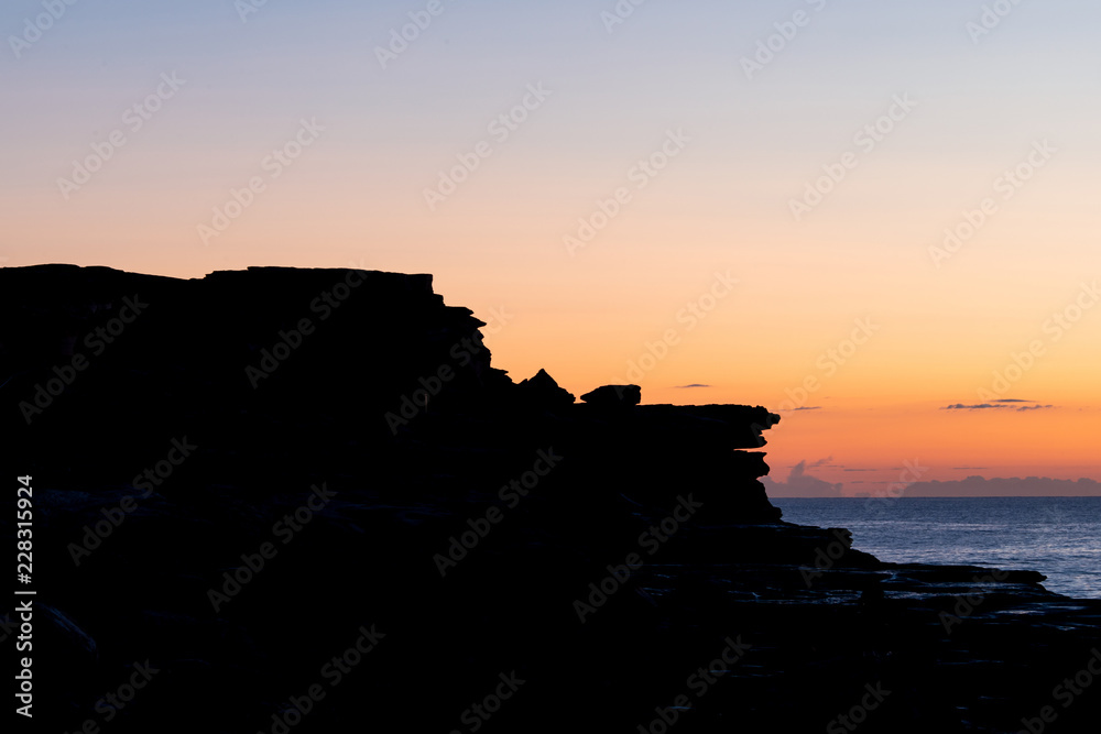 Silhouette of rock cliff with orange sunrise sky.